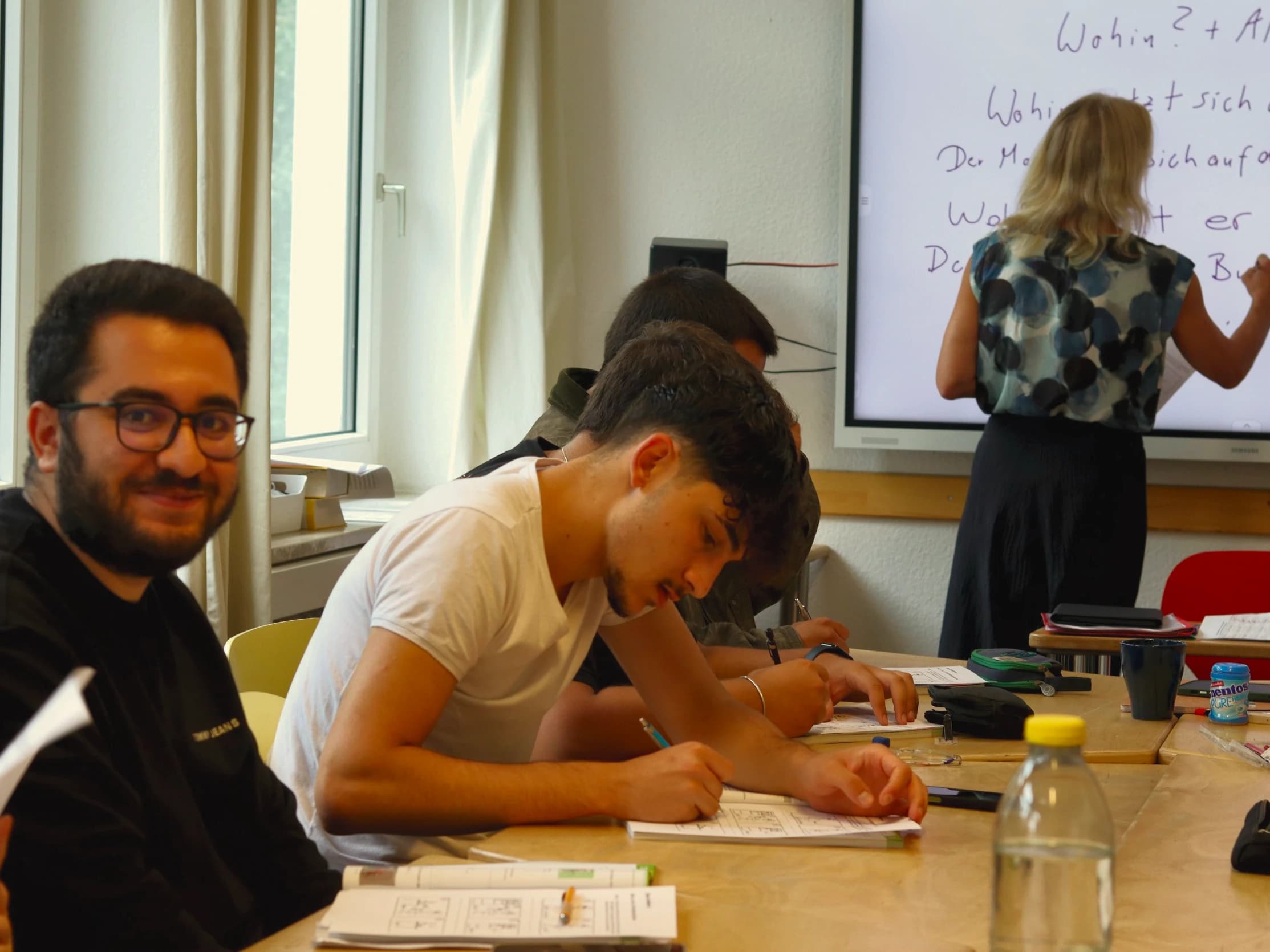 A learner smiles up from his exercise while the teacher writes grammar on the board